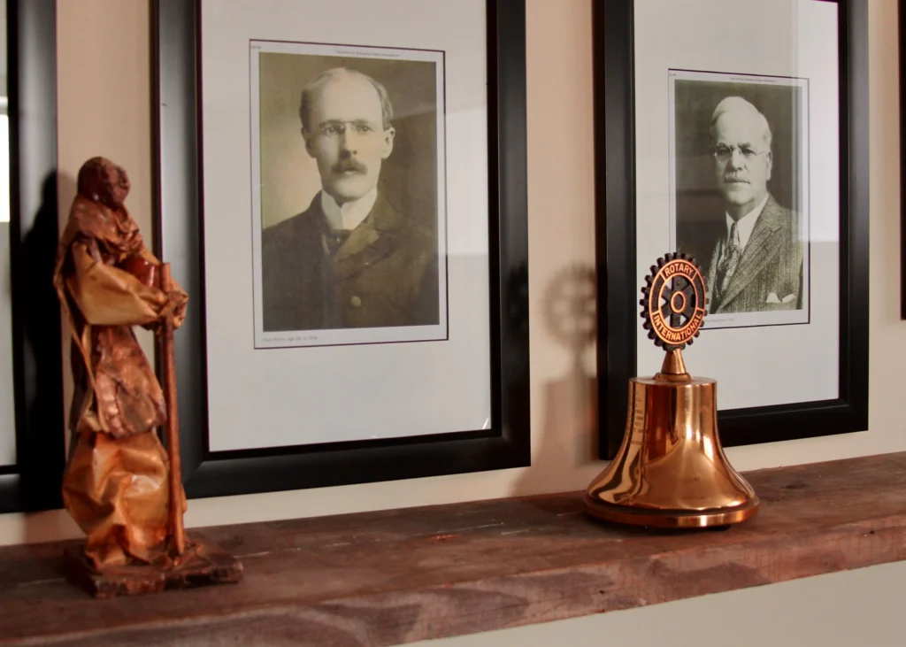 Rotary History Display – Detail Framed historic portraits, a carved figure, and a Rotary bell displayed on a shelf.
